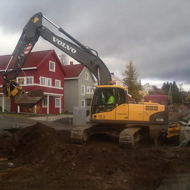 Gravemaskin arbeider ved rød bolig i et nabolag med mørke skyer og høstfarger i bakgrunnen.
