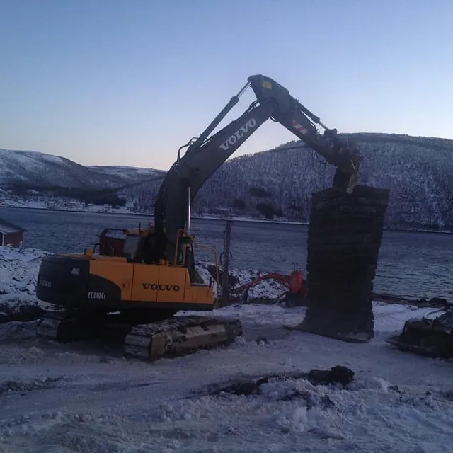 Gravemaskin løfter stablede matter på snødekt anleggsområde ved vann og fjell i vinterlys.