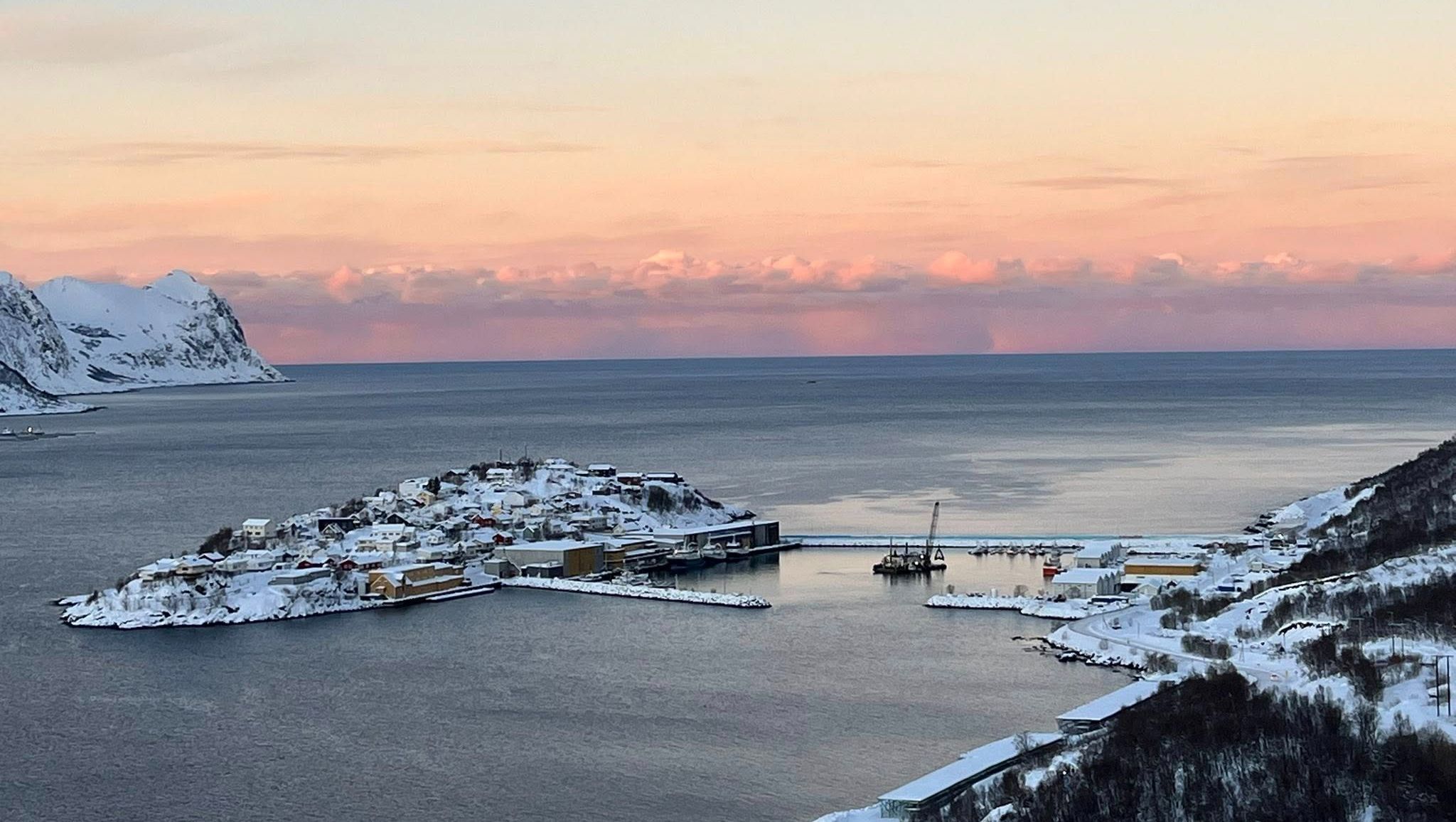 Havnemiljø ved kysten med flytekran, snødekte bygg og rosa kveldshimmel i horisonten.