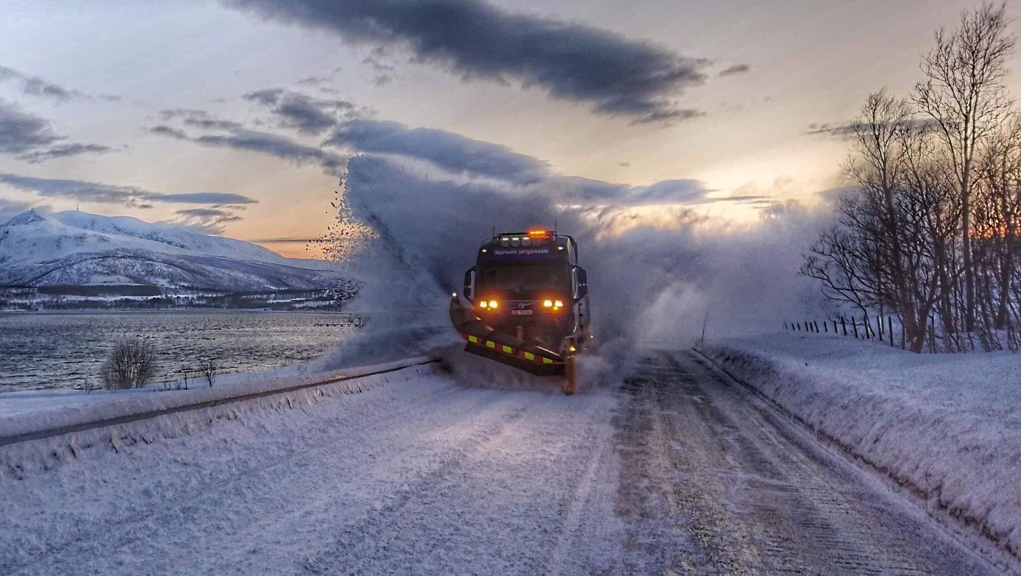 Brøytebil kaster snø høyt til siden mens den rydder vei i vinterlandskap ved sjøen.