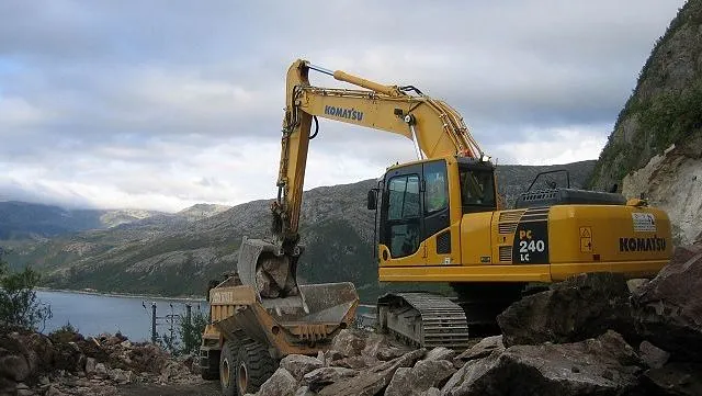 Veivals parkert på gruslagt område ved fjord, med snødekte fjell og trær i bakgrunnen.
