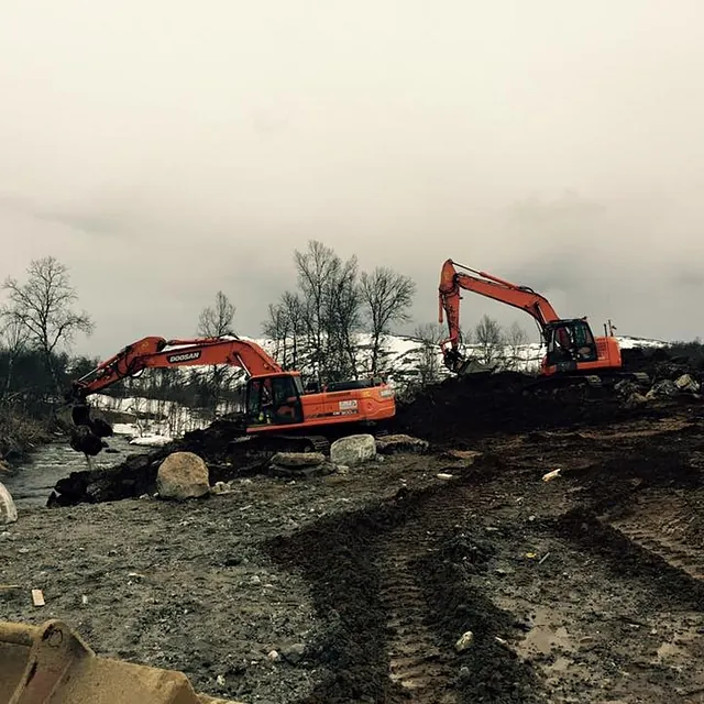 To gravemaskiner graver langs bekk i vinterlandskap med snø og nakne trær.