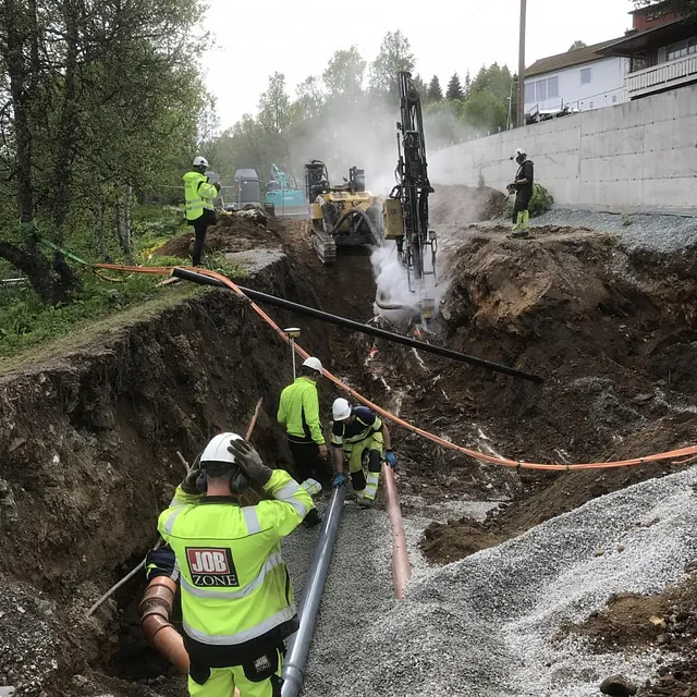Anleggsarbeidere jobber i dyp grøft mens borerigg avgir damp ved bolighus og trær.