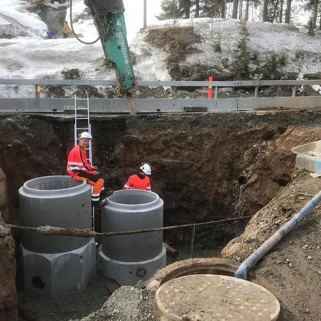 To anleggsarbeidere i grøft monterer store betongrør, med gravemaskin og snø i bakgrunnen.