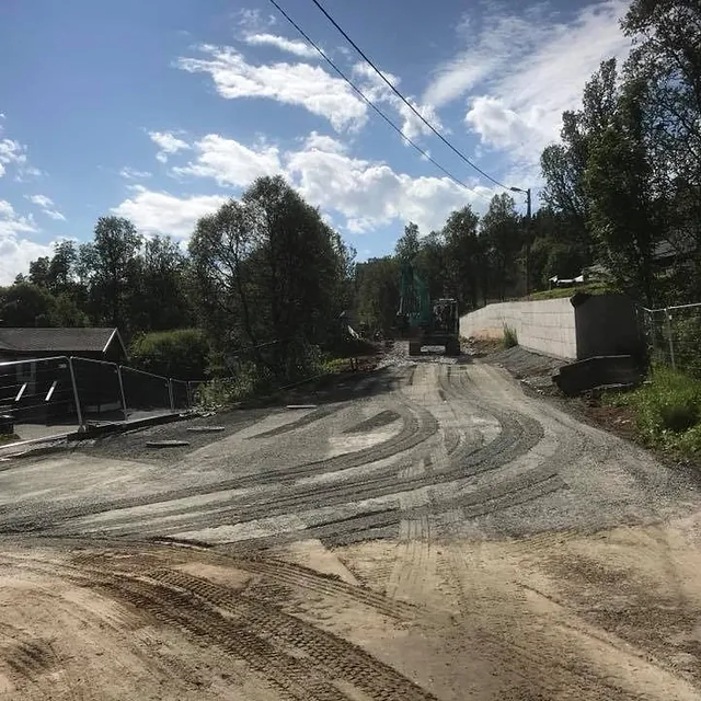 Gruslagt anleggsvei under arbeid mellom boliger og trær, med gravemaskin i enden av veien.