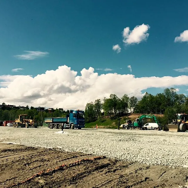 Maskiner og lastebil jobber på et flatt, gruslagt anleggsområde under blå himmel med skyer.