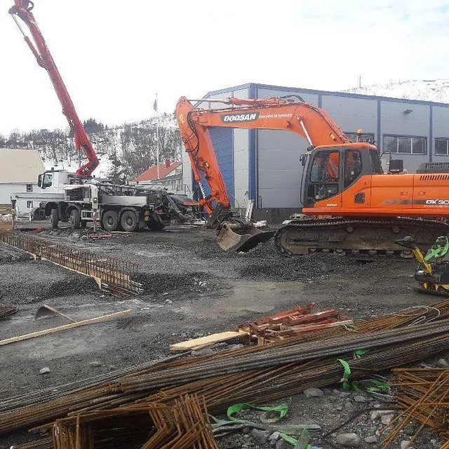 Gravemaskin og betongpumpebil i arbeid på en byggeplass foran et blått industribygg.