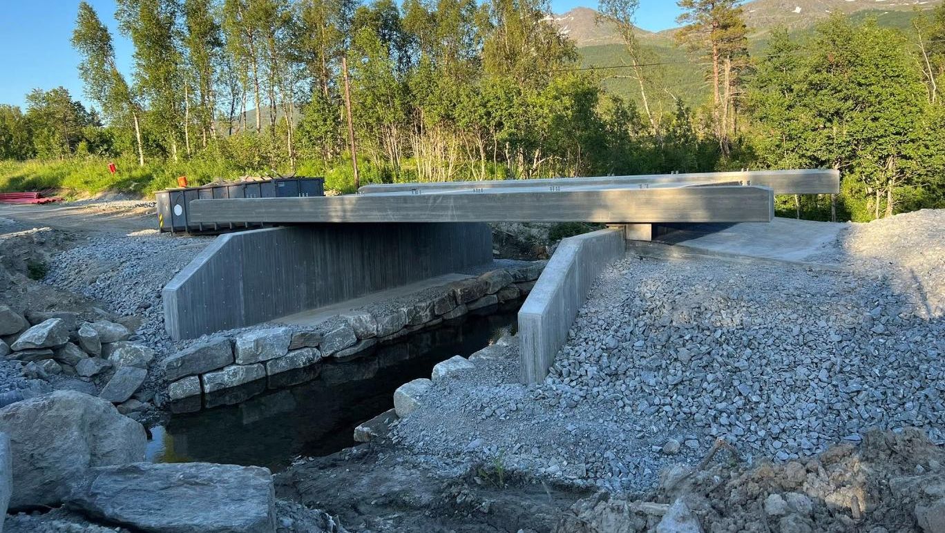 Elveforbygning med naturstein og støpt betongbru under montering i solfylt skogsområde.