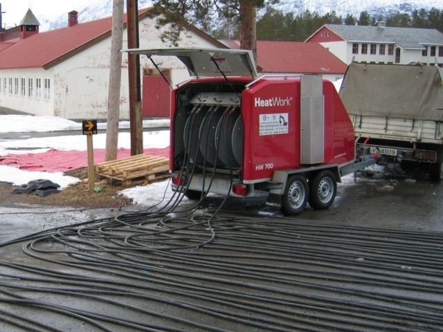 Varmeanlegg på tilhenger med mange varmeslanger rullet ut på bakken foran bygninger.