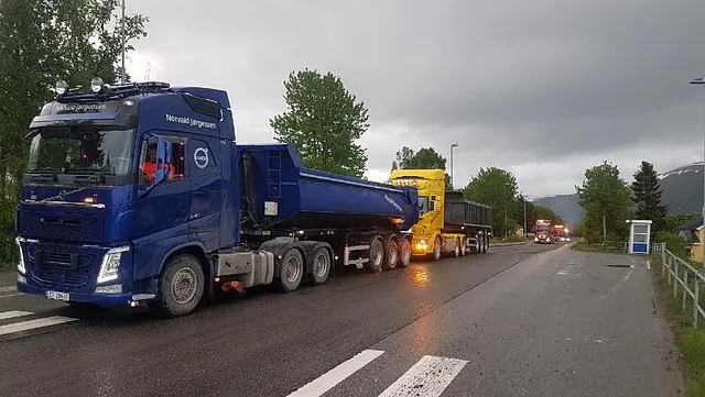 To lastebiler med hengere står parkert langs våt asfaltvei under mørke regnskyer.