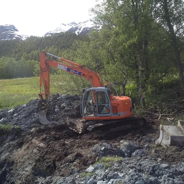 Gravemaskin med smalskuff jobber i skogkant med snødekte fjell i bakgrunnen.
