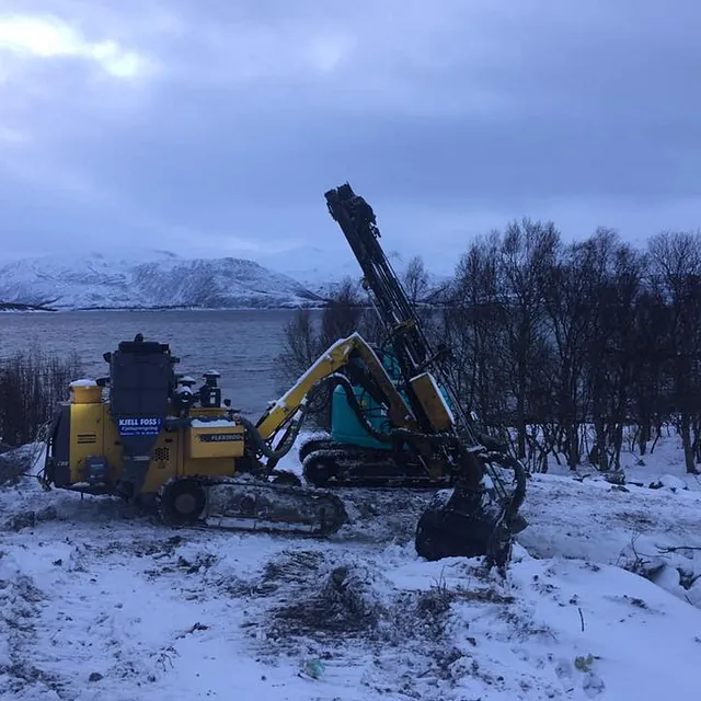 Borerigg og maskin arbeider i snødekt landskap ved vann, omgitt av trær og vinterfjell.