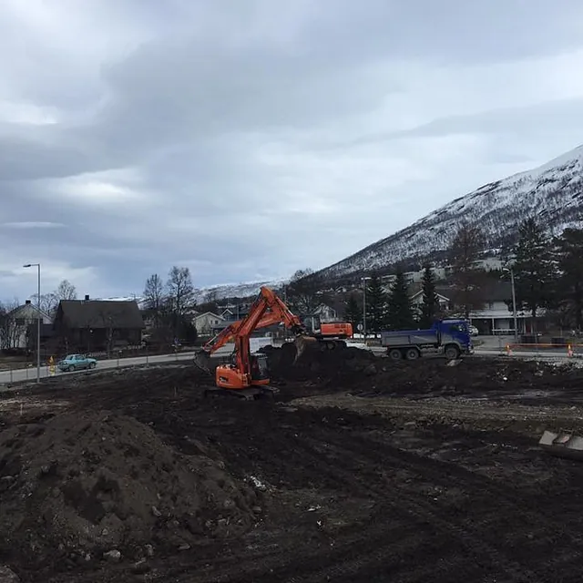 Gravemaskin laster masser i lastebil på byggeplass med fjell og boliger i bakgrunnen.