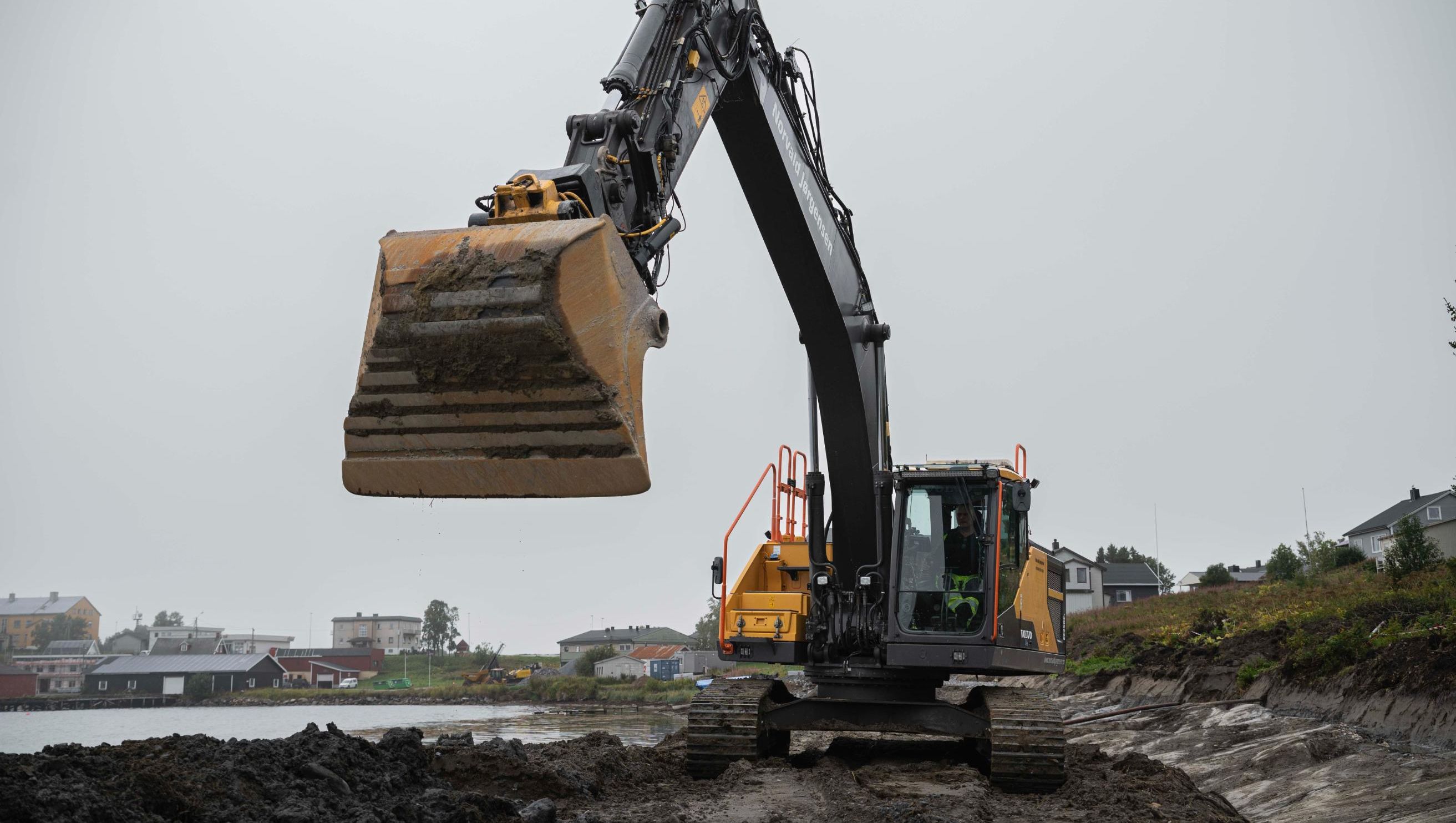 Gravemaskin med hevet skuff står ved vannkanten i et vått og gjørmete landskap.