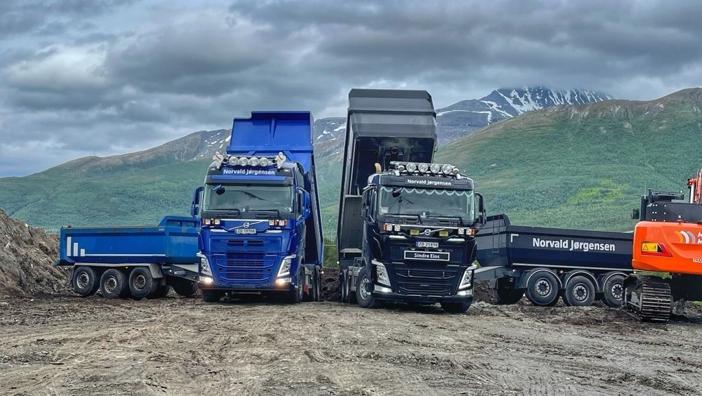 To lastebiler tipper masser på en anleggsplass foran grønne fjell og grå himmel.