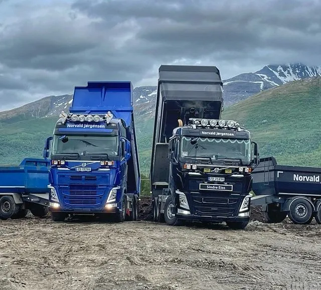 To lastebiler tipper masser på en anleggsplass foran grønne fjell og grå himmel.