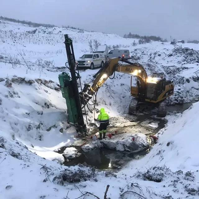 Gravemaskin og arbeider utfører boring i snødekt grøft, med servicebiler i bakgrunnen.