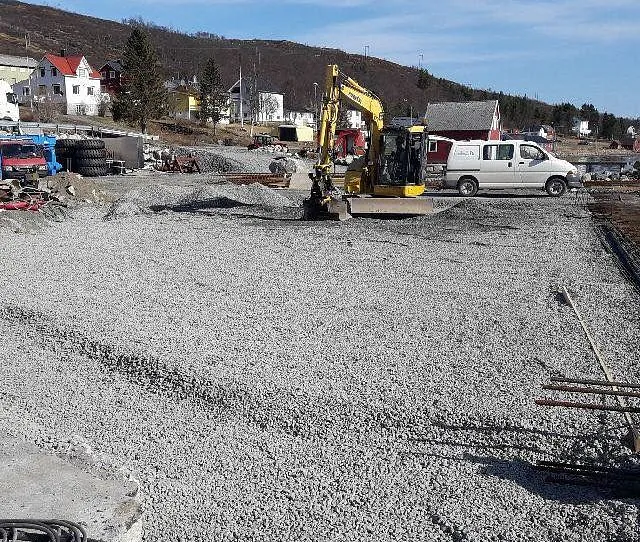 Anleggsområde med grusdekke, gravemaskin og hvit varebil foran bolighus og sjø.