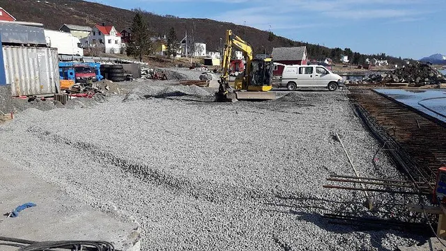 Anleggsområde med grusdekke, gravemaskin og hvit varebil foran bolighus og sjø.